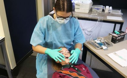 A woman in a blue gown, green gloves and facemask looks at the teeth of a child lying in front of her